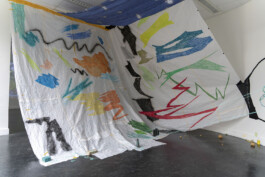A large drawing on paper hangs from the ceiling of a room and comes forward onto the room being held in place by 4 different bricks. The drawings is made up of colourful squiggles that dances and overlap with each other 