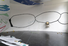 a room with a curved wall. On it hang two shelves, one with a tv and another one with a dvd player. On the wall behind the tv we also see a chain drawn with black tape. Hanging from the ceiling a series of strings attached to some bricks from which a sculpture made out of a chain hangs. 